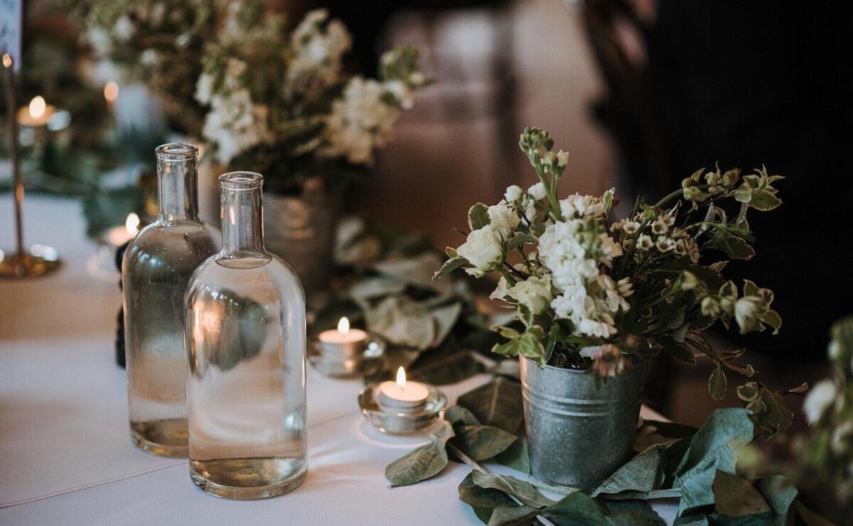 White flowers in a bucket, water bottles and candles on a table decorated with leaves