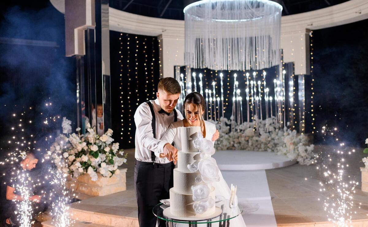 Front view happy married couple loving man and woman cutting big white cake on wedding ceremony at night Standing near welldecorated luxury altar with flowers and sparklers Special event