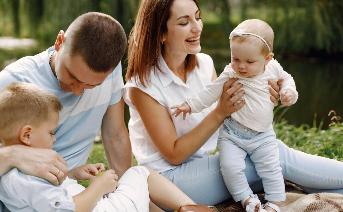 Mother, father, older son and little baby daughter sitting on a picnic rug in the park. Family wearing white and light blue clothes